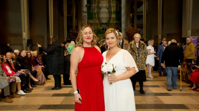 A woman in a red dress stood next to a woman in a white wedding dress stood in front of the large tapestry in Coventry Cathedral