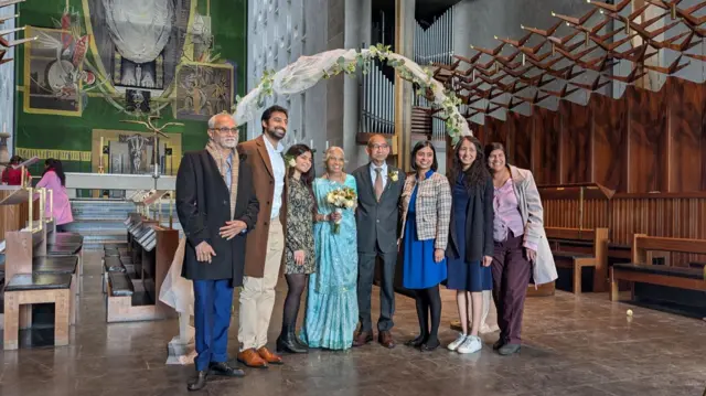 A group of eight people, one in a saree and others in suits and dresses, stood in front of a flora arch in Coventry Cathedral