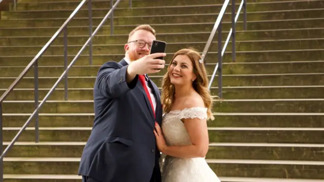 A man wearing glasses in a dark suit and a red tie next to a woman in a white wedding dress in front of the steps at Coventry Cathedral