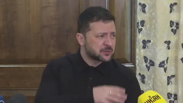 Zelensky sitting down in front of wooden door and curtain as he addresses reporters from inside a hotel in Munich