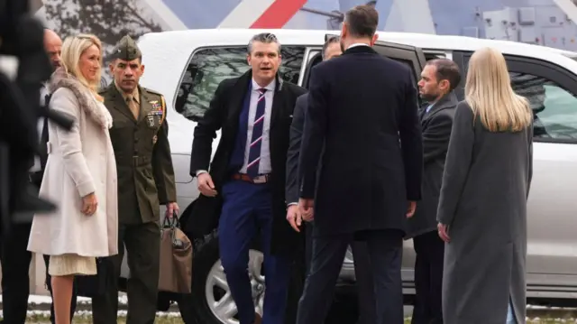 Pete Hegseth steps out of a white car as he arrives in Warsaw, Poland and is welcomed by local officials. A man in green uniform stands to his right holding a brown back, Hegseth's wife Samantha in a fur-collared coat to his right