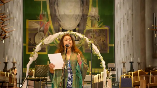 Yasmin Wilde reading from a sheet in front a large green tapestry in Coventry Cathedral