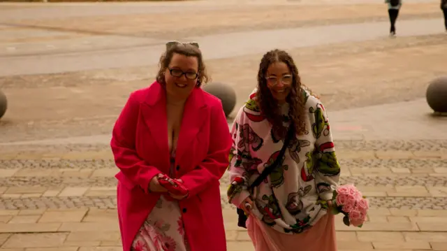 A woman in a red coat and a floral dress next to a woman in a Shrek-themed jumper, walking up some steps