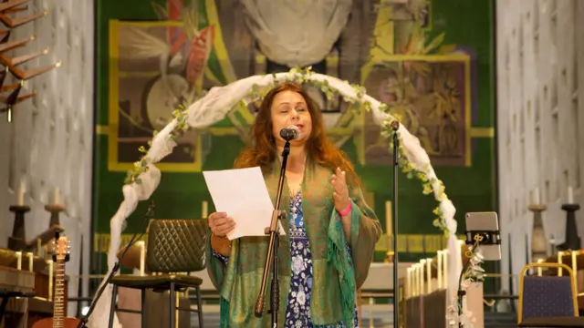 Actress Yasmin Wilde reading from a sheet in front of a green large tapestry