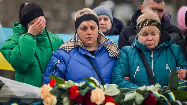 Natalya is flanked by two other women at her husband's funeral, with flowers visible in the foreground