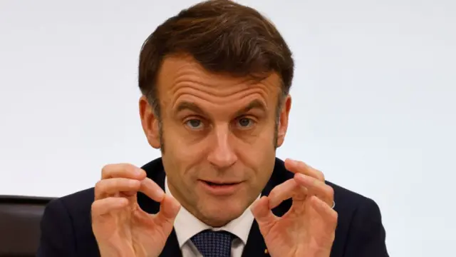 Emmanuel Macron with his hands in front of him gesturing as he speaks sitting down in chair. He's wearing a dark blue suit, white shirt and blue tie, a white wall in the background