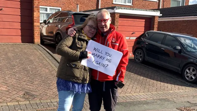 Tracey Draper, holding paper saying 'Will you marry me... again?' next to Kevin, dressing in a red Royal Mail jacket and black trousers on a driveway in front of a red brick house