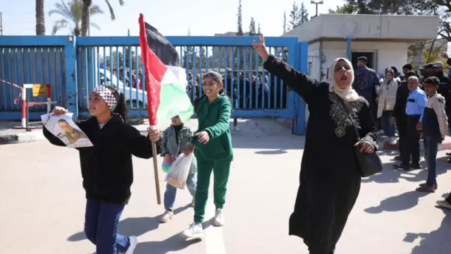 A women and three young children celebrate in the street, one child holding a Palestinian flag and a piece of paper with a man's face on it