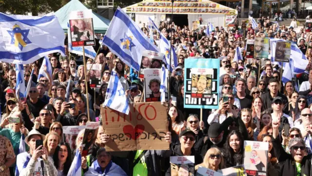 A crowd watches outside, holding Israeli flags and images of the hostages