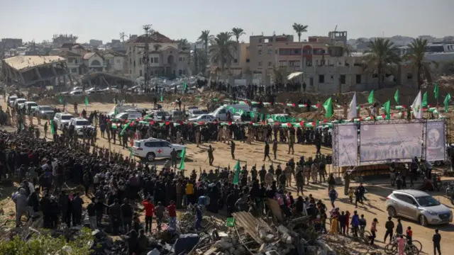 Red Cross vehicles enter a crowded area, with a clearing in the middle where the stage is set for the handover