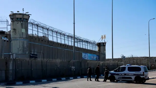 A police car and three people stand outside a prison which has high walls and barbed wire