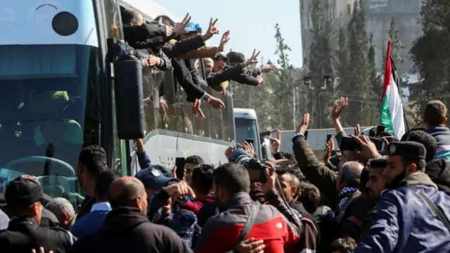 A Palestinian flag among a crowd of people as buses arrive with men leaning out the windows