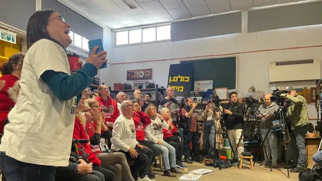 People watch a screen together, with media at the side filming them