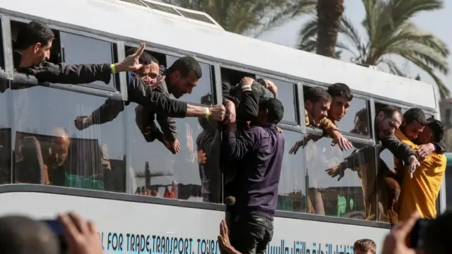 Men hang out of the windows of a bus. Some men in the surrounding crowd have been lifted up and are hugging those leaning out of the bus windows