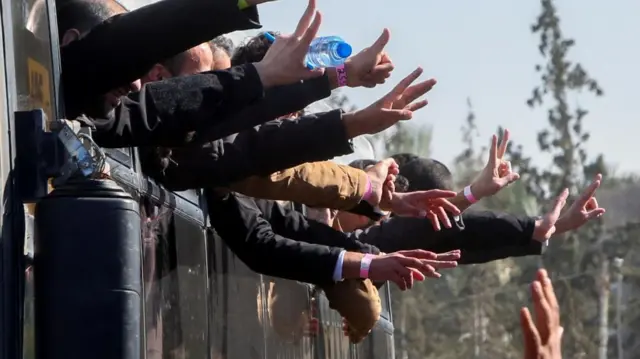 Men hang out of bus windows and make gesture with their hands, many forming a 'V' shape with two fingers