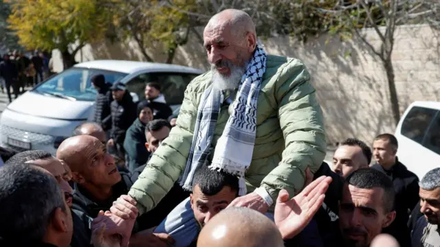 A man with a white and grey beard sits on the shoulders of a younger man as people crowd around