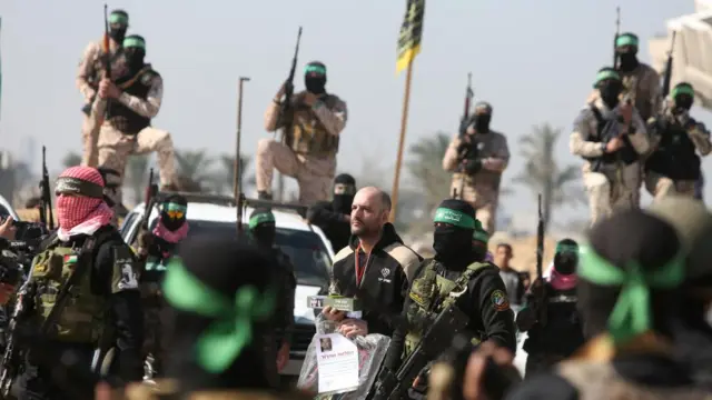 A man walks surrounded by armed men with their faces covered