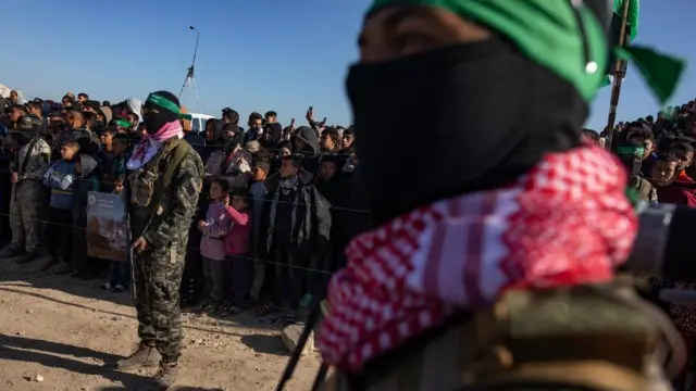 Palestinians stand next to fighters from the al-Qassam Brigades prior to a handover ceremony of three Israeli hostages to the Red Cross