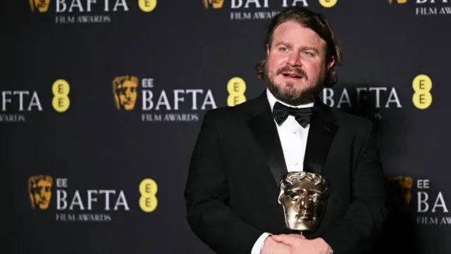 Brady Corbet, a man with long brown hair and facial hair wearing a black tuxedo, holds his BAFTA on a red carpet in front of a black wall with the BAFTA and EE logos.