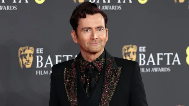 David Tennant walks on Bafta red carpet, wearing a black blazer and shirt with embroidered lapels and collars.