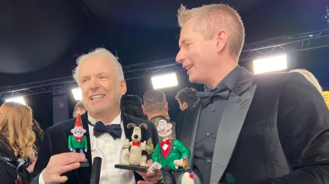 Nick Park and Merlin Crossingham hold Wallace & Gromit figurines on red carpet, wearing black tuxedos.