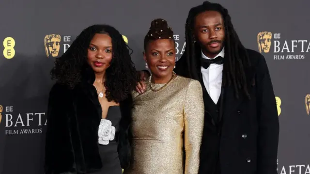 Angela Patton wears a gold gown and gold necklace. She smiles with two people on either side of her.