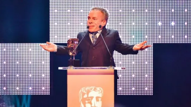 Actor, comedian and presenter Warwick Davis standing on the Bafta stage with his arms outstretched, accepting an award. He is wearing a suit and tie and stands in front of a glittery stage background