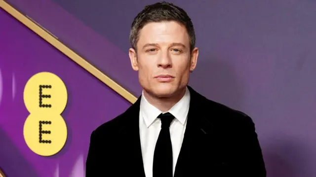 James Norton wears a black suit in front of a purple backdrop.