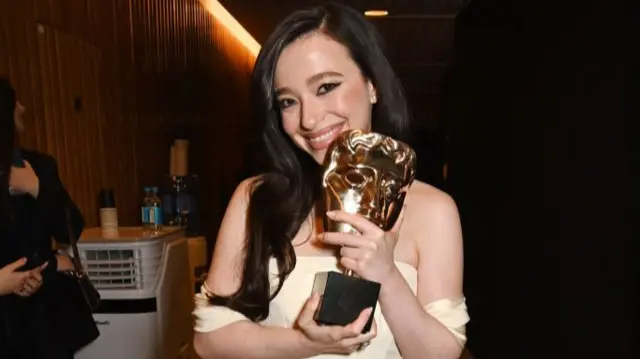 Mikey Madison, who has long brown hair and wears a sleeveless yellow dress, holds her BAFTA and smiles.