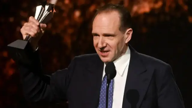 Ralph Fiennes at the 30th Annual Critics Choice Awards held at The Barker Hangar on February 7, 2025 in Santa Monica, California. He is holding aloft the silver award and he is wearing a navy suit, white shirt and blue and white polka dot tie.