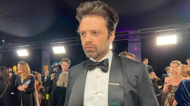 Sebastian Stan wears a black tuxedo with a white shirt on the red carpet.