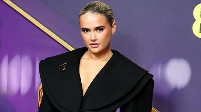 Molly Mae wears a black blazer with exaggerated shoulders. A purple Bafta backdrop is in the background.