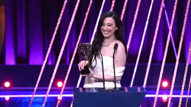 Mikey Madison, who has long brown hair and wears a sleeveless yellow gown, holds her BAFTA on stage in front of a podium.