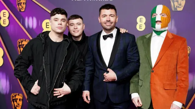 A group picture of the Kneecap cast, one wearing the colours of the Ireland flag on a face mask and a green and orange blazer.