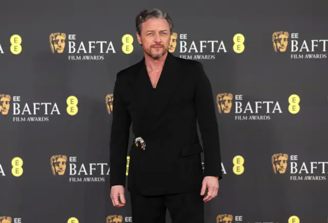 James McAvoy in a black suit standing in front of a background with the Bafta logos.