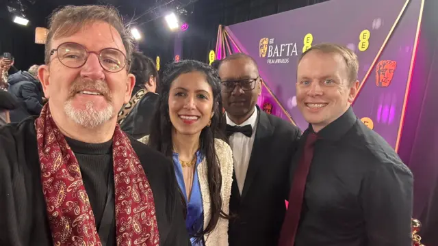 BBC journalists smile with on the red carpet with a purple sign of the Bafta awards in the background