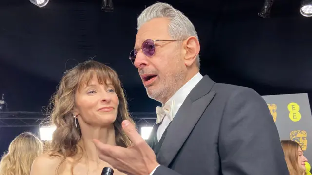 Jeff Goldblum speaks beside his wife. He wears a black suit with white bow tie and round purple glasses.