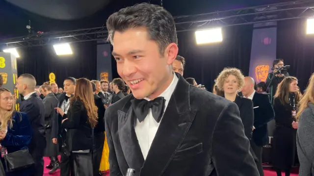 Henry Golding wears a black tux and bow tie on the red carpet.