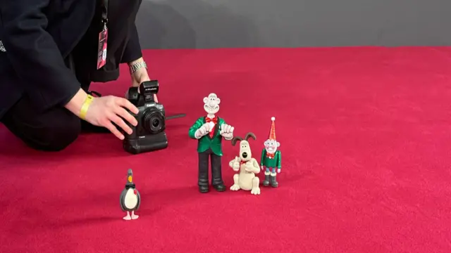 Figurines of Wallace and Gromit are placed on the red carpet as someone holds a camera in the background.