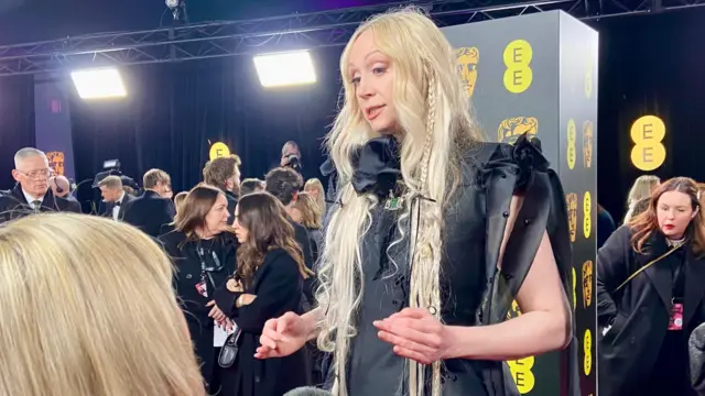 Gwendoline Christie wears a black outfit with long blonde hair. Several people are in the background, some holding cameras.