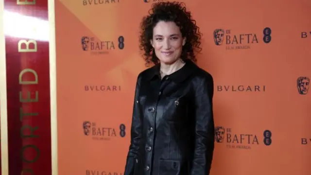 Coralie Fargeat wears a black leather shirt with black trousers. There is an orange background with the Bafta logo in black written over it.