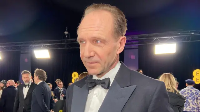 Ralph Fiennes on the red carpet wears a suit and bow tie.