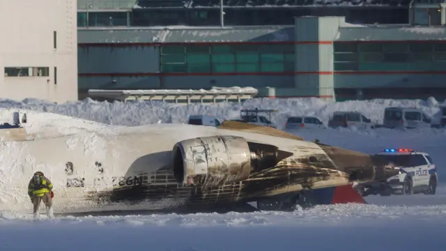 The image shows the plane flipped upside down on a tarmac covered in snow.