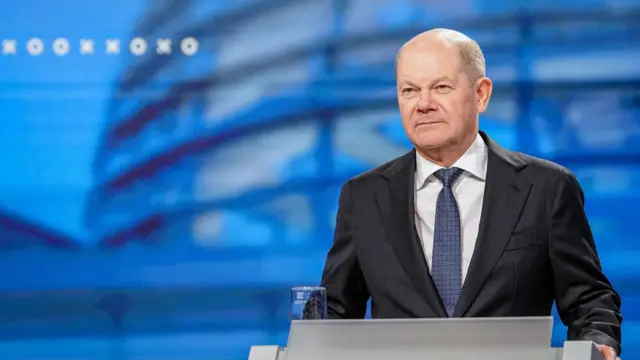 German Chancellor Olaf Scholz standing at a podium against a blue backdrop.