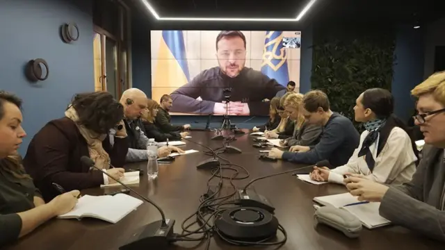 Zelensky on a video screen behind journalists sat at a long table