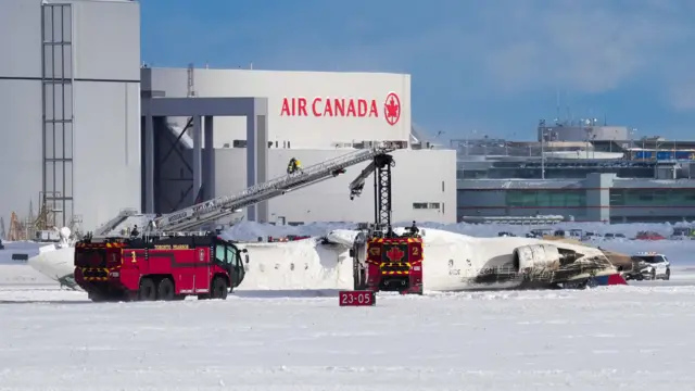 A plane flipped over laying on a runway with snow and firetrucks around it