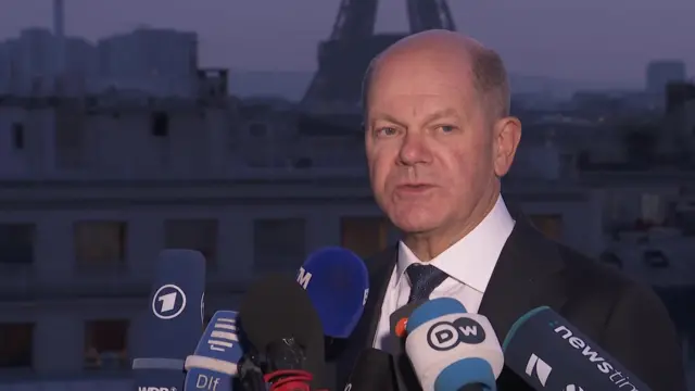 Olaf Scholz speaking to journalists from a rooftop position, with the Eiffel Tower in the background.