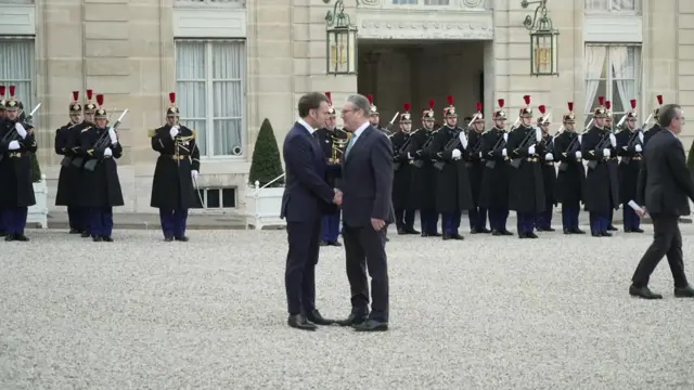 Starmer and Macron shake hands in the Elysee Palace courtyard - people in ceremonial military uniforms stand behind them