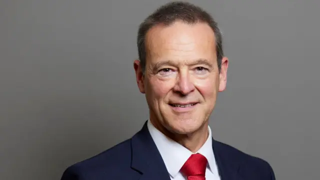 Headshot of Lord Simon McDonald wearing a blazer, white shirt and red tie.