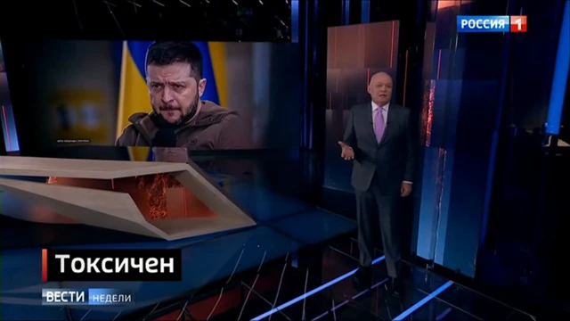 A screenshot of a TV show with a presenter stood wearing a suit in a TV studio with a picture of Zelensky on a big screen behind him